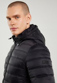 Hombre con cabello corto que lleva una chaqueta negra acolchada con capucha, cremallera y cordones ajustables sobre un fondo claro.