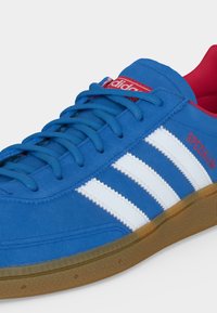 Μπλε παπούτσι Adidas Spezial με λευκές ρίγες, κόκκινες λεπτομέρειες λογότυπου, μπλε κορδόνια και καφέ λαστιχένια σόλα.