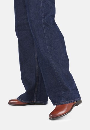 Jean en denim bleu foncé évasé avec des coutures visibles et une texture légèrement rugueuse, associé à des bottes en cuir marron arrivant à la cheville avec un bout pointu.