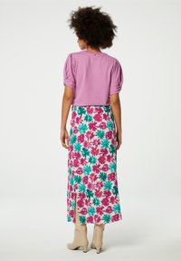 Lila Bluse mit Puffärmeln kombiniert mit einem floral gemusterten Midirock in Pink- und Türkis-Tönen. Das Modell trägt beige Knöchelstiefel. Helle, strukturierte Materialien.