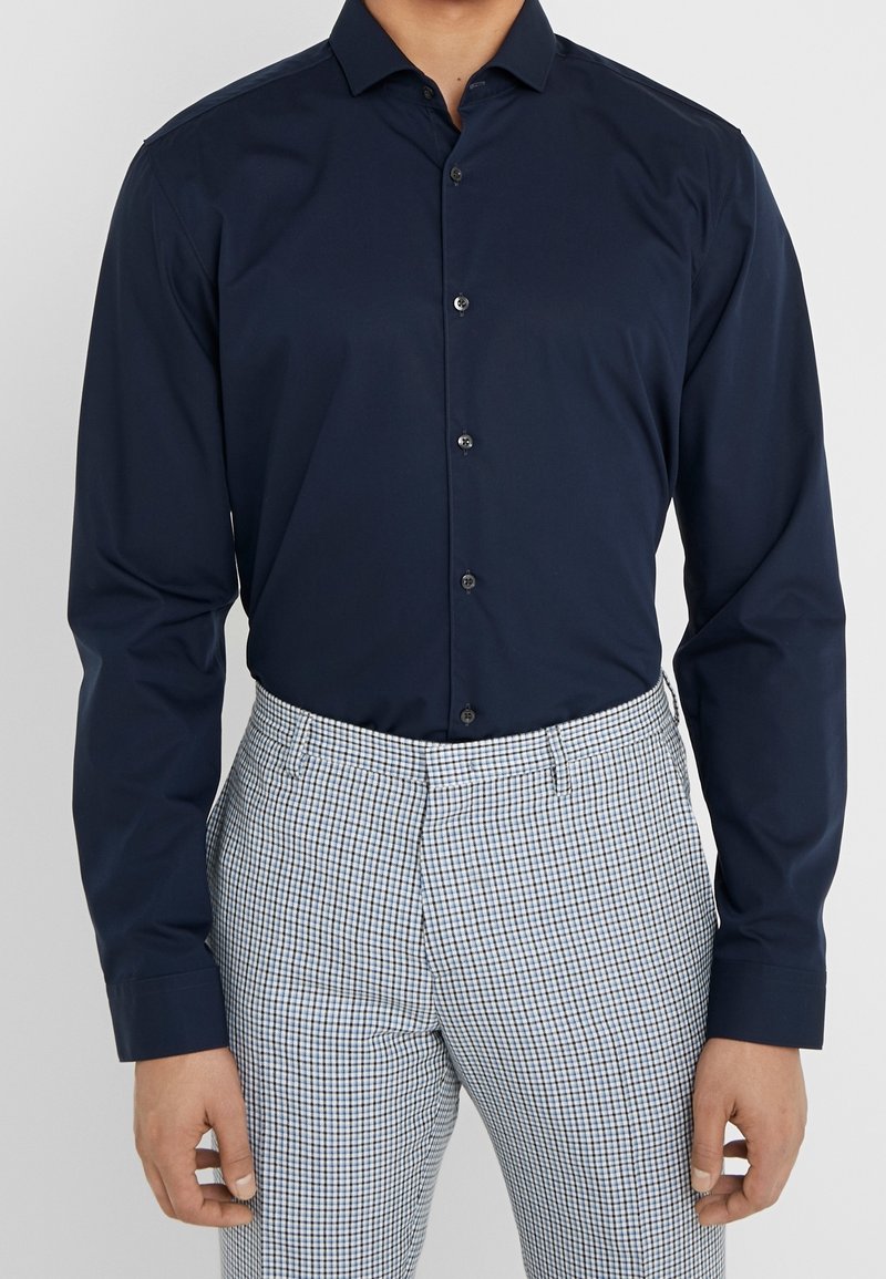 Camisa de botones en azul marino con cuello, de manga larga y corte regular; combinada con pantalones de cuadros a medida en tonos azul y blanco.