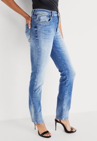 Lichtblauwe denimspijkerbroek met een rechte pasvorm, vijf zakken, subtiele gebruikssporen en een normale taille. Draagt met zwarte strappy hakken.