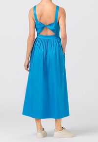 En blå bomullsklänning med en figurnära byst, breda axelremmar, en urskuren rygg med en rosett och en veckad, medellång kjol. Bärs med ljusa sneakers.