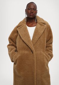 Tannenkleurige, fuzzy textuur jas met een sjaalkraag, voorzakken en een enkele knoopsluiting. Gelaagd over een witte top.