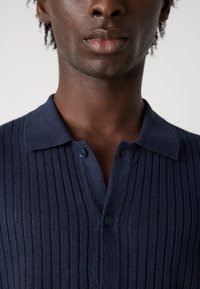 Polo shirt in maglia blu navale con righe verticali, colletto e bottoni. Il tessuto sembra leggero e testurizzato.