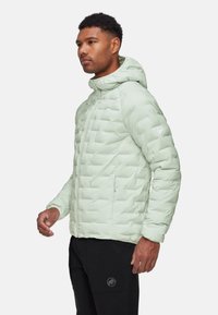 Hellgrüne Steppjacke mit Kapuze, versehen mit einem gesteppte Design und einem vorderen Reißverschluss, kombiniert mit schwarzen Hosen. Logo auf dem Ärmel.