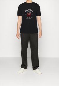 Černé tričko s červeným a bílým logem "HILFIGER" a textem "New York", spárované s tmavými kalhotami a bílými teniskami. Jednoduchý design s kulatým výstřihem.