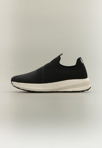 Μαύρο αθλητικό παπούτσι χωρίς κορδόνια με πάνω μέρος από υφαντό ύφασμα με υφή και λευκή μαλακή σόλα, χαμηλό κόψιμο με θηλιές στο γλώσσα και τη φτέρνα.