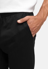 Schwarze Sporthose aus strukturiertem Stoff, mit einer Seitentasche und einer Nahaufnahme einer Hand, die in der Tasche liegt.