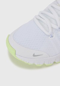 Hvide sportsko med en tekstureret overdel, lys grøn gummisål og sølvfarvet Nike-logo på siden, der har hvide snørebånd og mesh-detaljer.