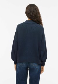 Maglione blu navy a coste con collo ampio e maniche a palloncino, abbinato a jeans di denim blu, mostrato di spalle.