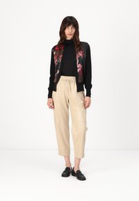 Zwarte cardigan met bloemenprint aan de voorkant, lange mouwen en knoopsluiting, gecombineerd met beige cropped broeken en zwarte instappers.