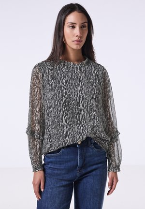 Blouse transparente à motifs noir et blanc avec des manches longues, des poignets ruffles et un col rond, associée à un jean en denim bleu.