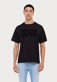 Schwarzes Baumwoll-T-Shirt mit dem auffälligen Text "VERSACE JEANS COUTURE" in einer geschichteten Schriftart. Kurze Ärmel und runder Ausschnitt.