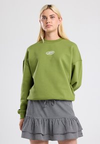 Pullover verde oversize con scollatura rotonda e logo ricamato sul petto, abbinato a una gonna a balze in bianco e nero a quadri.
