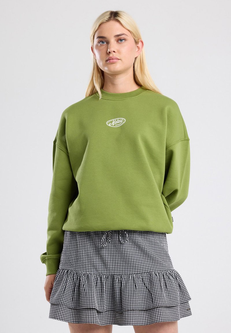 Pullover verde oversize con scollatura rotonda e logo ricamato sul petto, abbinato a una gonna a balze in bianco e nero a quadri.