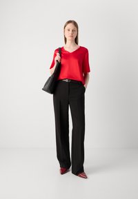 Top rosso di raso con scollatura a V, abbinato a pantaloni neri a gamba larga e scarpe rosse a punta. La modella tiene una borsa a tracolla nera.