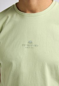 Camiseta de algodón verde claro con cuello redondo y un logotipo impreso "PETROL IND MIAMI, FL. U.S.A." en un color contrastante.