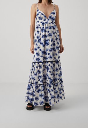 Robe longue blanche sans manches avec imprimé floral bleu ; encolure en V, jupe à volants et bordure noire. Associée à des sandales à plateforme noires.