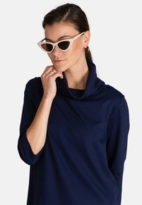 Blusa navy de cuello vuelto con mangas hasta los codos, combinada con gafas de sol de forma de gato blancas con lentes oscuros. Textura de tela suave y lisa.