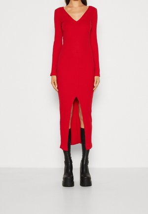 Kobieta nosząca czerwoną, prążkowaną sukienkę z dzianiny z długim rękawem i rozcięciem z przodu, w połączeniu z czarnymi masywnymi butami na platformie.