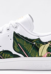 Valkoinen nahkainen tennari, jossa on vihreitä ja vaaleanpunaisia kirjailtuja lehtikuvioita, pyöreät nauhat ja valkoinen kumipohja, jossa hillitty logodetaili.