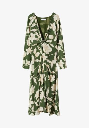 Femme portant une robe midi verte à imprimé floral blanc, manches longues, détail noué à l'avant, et sandales à talons beiges, debout sur un sol uni.