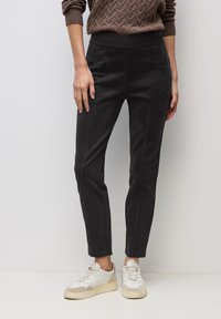 Persona vistiendo pantalones negros ajustados, suéter marrón con textura y zapatillas blancas, de pie contra un fondo blanco liso.