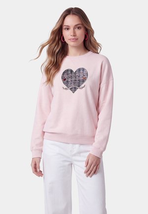 Donna con lunghi capelli mossi indossa una felpa rosa con un cuore a quadri e testo, abbinata a pantaloni bianchi e orecchini a cerchio rosa.