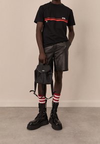 Camiseta negra de manga corta con una franja roja, pantalones cortos de cuero negros, mochila negra y botas de combate negras robustas con calcetines a rayas rojas y blancas.