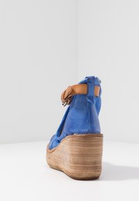 Modré semišové klínové sandály s hnědým kotníkovým páskem a dřevěnou platformou na bílém povrchu proti bílému pozadí.