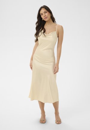 Femeie cu păr lung și închis la culoare purtând o rochie midi crem fără mâneci și pantofi cu toc deschis bej, stând în fața unui fundal simplu.