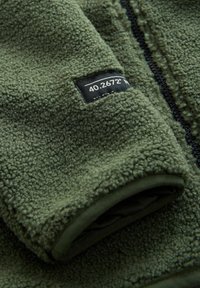 Fleece jacket in dark green with a textured surface. Features a zippered front and a black fabric label on the sleeve.
