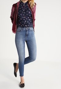 Cazadora bomber de satén marrón sobre una camisa de botones oscura con flores, combinada con jeans skinny azul claro y bailarinas negras con acentos de lazo.