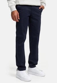 Pantalones chinos azul marino hechos de una suave tela de algodón, que presentan un corte cónico clásico y bolsillos laterales, combinados con zapatillas blancas.