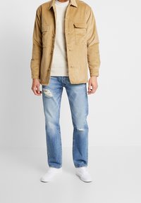 Kordjacka i ljusbrun, med två bröstfickor, kombinerad med slitna ljusblå jeans och vita sneakers.