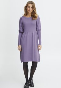 Långärmad lila klänning i mjukt tyg, med rund halsringning, samlad midja och lös ärmar. Kombineras med svarta strumpbyxor och loafers.
