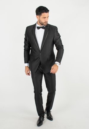 Tuxedo noir avec revers en satin, coupe ajustée et fermeture à bouton unique. Chemise blanche en dessous, assortie à un nœud papillon noir et des chaussures en cuir.
