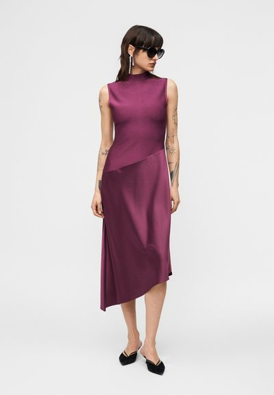 Γυναίκα που φοράει ένα μωβ ασύμμετρο μίντι φόρεμα χωρίς μανίκια, μαύρα μυτερά παπούτσια, μεγάλα σκουλαρίκια και μαύρα γυαλιά ηλίου, στέκεται σε λευκό φόντο.