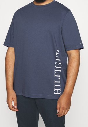 Man draagt een marineblauw T-shirt met korte mouwen en de tekst "HILFIGER" verticaal gedrukt aan de linkerkant, gecombineerd met een donkere broek.