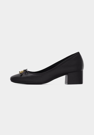 Black leather low-heeled pump with rounded toe, small bow, and gold circular emblem on the front.