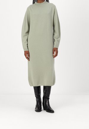 Kobieta ubrana w długą, jasnooliwkową dzianinową sukienkę w połączeniu z czarnymi skórzanymi kozakami za kolano, stoi na białym tle.