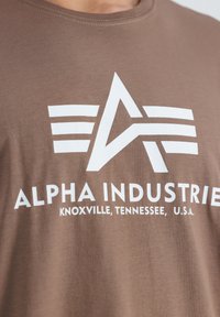Camiseta de algodón marrón con un logo gráfico blanco que presenta letras estilizadas y rayas, tejido texturizado y cuello redondo.