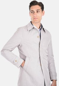 Trench coat grigio chiaro, resistente all'acqua, con colletto a punta, chiusura frontale con bottoni, tasche laterali e cuciture tono su tono. Texture liscia.