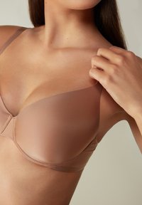 Beige podprsenka s kosticemi vyrobená z hladké, strukturované látky. Nastavitelné ramínka a švy po obvodu košíčků pro podporu a dokonalé přizpůsobení.