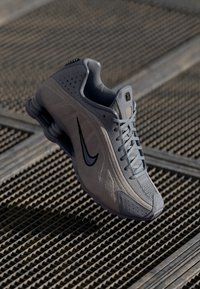 Pilka Nike sneakeris su zomšo ir tinklo viršumi, tekstūruotais detalėmis, akcentuotu swoosh logotipu ir matomu raištelių sistemos dizainu. Išryškintas šoko sole dizainas.