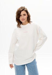 Cremefarbener Pullover mit langen Ärmeln, Rundhalsausschnitt und pinkfarbenem, gesticktem Schriftzug "MIRONS" mittig auf der Brust; kombiniert mit hellblauen Jeans.