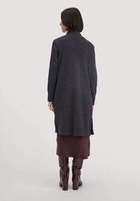 Langer grauer Cardigan mit hohem Kragen und seitlichen Schlitzen, aus strukturiertem Stoff. Getragen über einem bordeauxfarbenen Rock und kniehohen braunen Stiefeln.