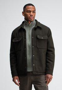 Dark green wool jacket with buttoned front, two chest pockets, and a collared neckline worn over a light grey zip-up sweater.