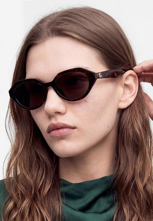 Jeune femme aux longs cheveux bruns portant des lunettes de soleil sombres et angulaires et un top vert foncé, ajustant la branche des lunettes.